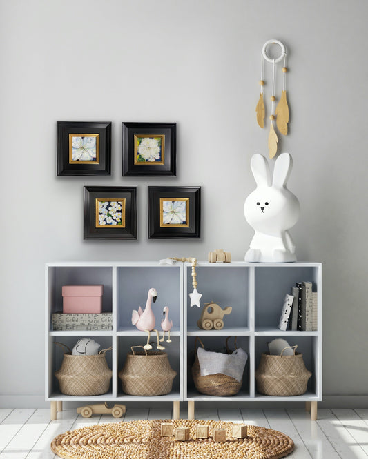 Four small floral paintings in black and gold frames arranged above white cubby shelves in a nursery with toys and woven baskets.