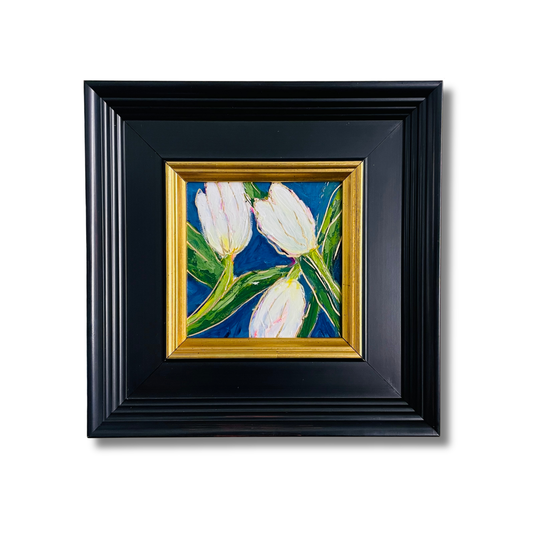 A framed painting of three white tulips with green leaves on a deep blue background, set in a gold inner frame and a larger black outer frame, shown against a plain white background.