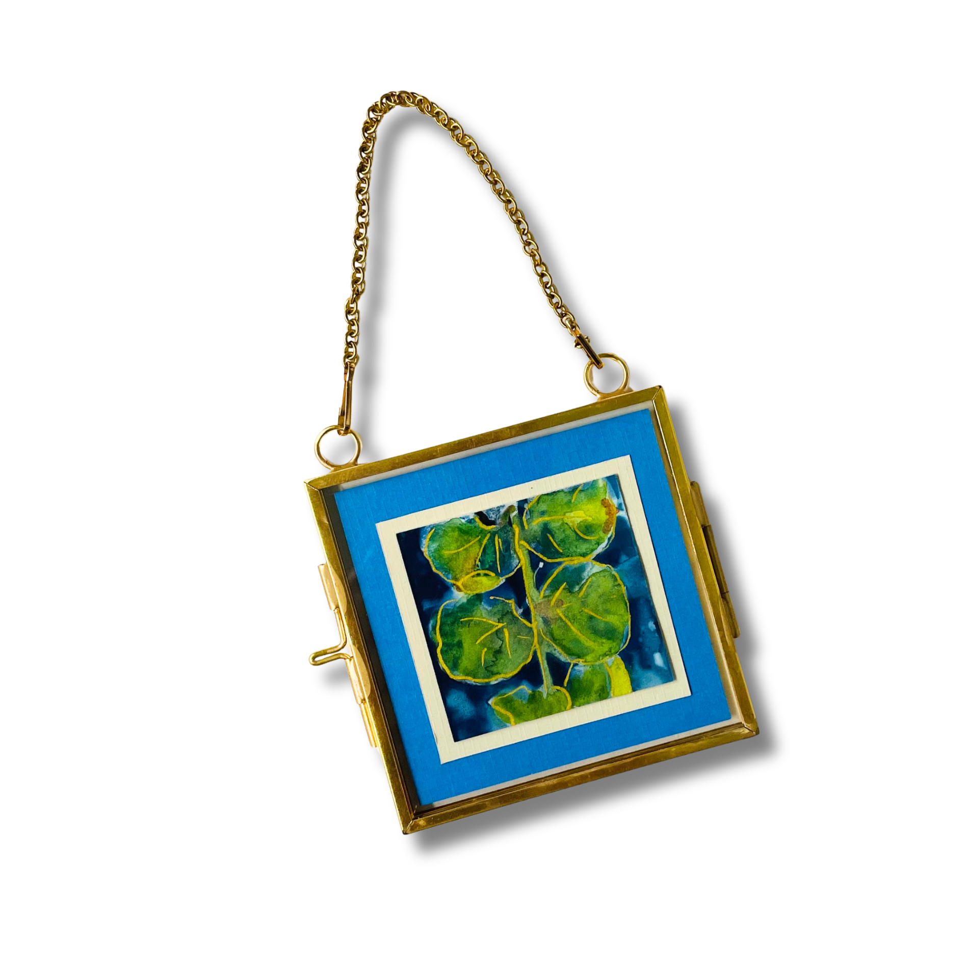 A square gold-framed hanging wall art piece with a vivid blue border and a central botanical painting of green leaves on a dark background, suspended by a gold chain against a white studio background.