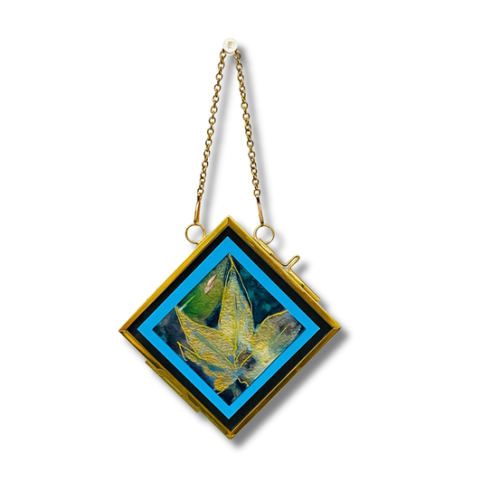 A diamond-shaped gold pendant with a hanging chain, featuring a central artwork of a green leaf on a blue and black background.