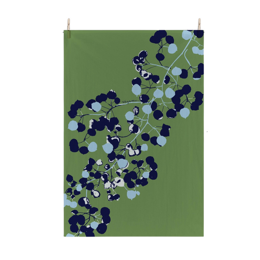 A rectangular green flour sack tea towel with a stylized botanical pattern of dark navy, light blue, and white aspen leaves, hanging from two small wooden clips against a plain white background.