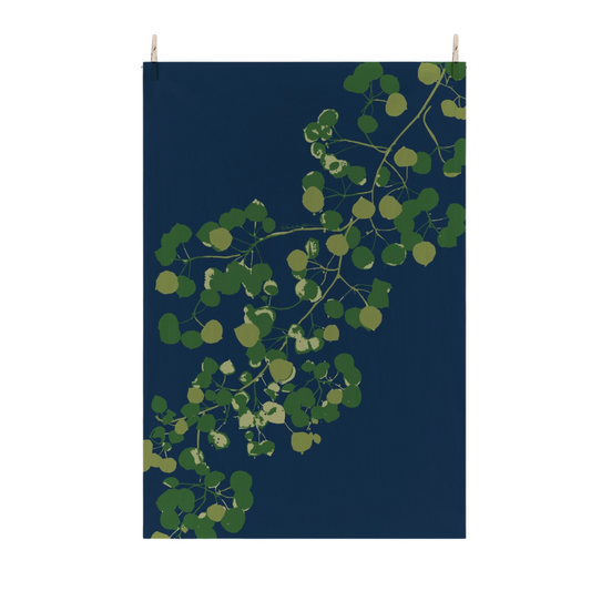 A vertical flour sack tea towel with a dark navy blue background and a stylized green aspen leaves, suspended by two small wooden pegs against a plain white wall.