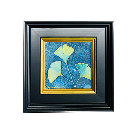 Framed square wall art featuring three yellow-green ginkgo leaves on a textured blue background, set within a gold inner frame and a black outer frame, shown against a plain white background.