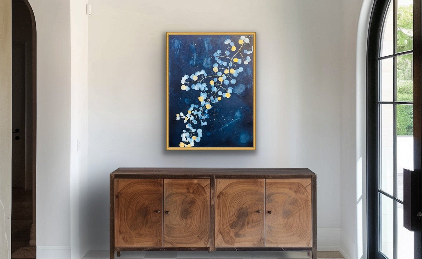 A framed abstract painting with deep blue tones and white floral elements hangs on a white wall above a wooden sideboard in a bright entryway with a checkered tile floor and arched doorway.