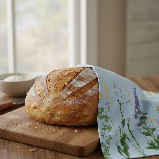 A round loaf of rustic bread sits on a wooden cutting board with a light blue floral cloth draped over the right side, set in a softly lit kitchen near a window.